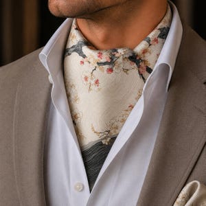 May include: An ascot featuring a cream-colored background with a delicate pattern of dark gray tree branches and pink and white cherry blossoms. The fabric displays a subtle wavy texture, worn tucked into a white collared shirt under a tan blazer.