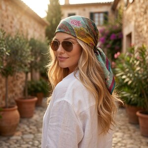 May include: A woman wearing a silk headscarf with a floral and geometric pattern in shades of teal, pink, and gold. She is also wearing gold-rimmed sunglasses and a white linen shirt. The background shows a cobblestone street and buildings.