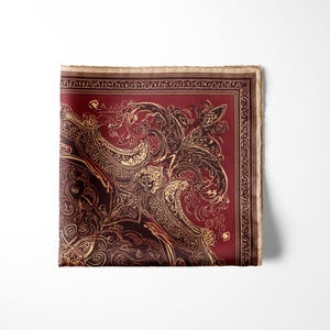 May include: A folded square pocket square with a burgundy background and ornate gold paisley design. The edges are bordered with a lighter tan color and a repeating pattern. The pocket square is made of a silky material.