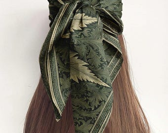 May include: A dark green headscarf with a repeating gold leaf pattern. The scarf is tied in a knot at the top of the head, with the ends hanging down. The scarf is made of a silky material.