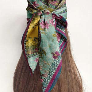 May include: A floral patterned headscarf tied around the head. The scarf features a light blue background with pink flowers and a yellow accent. The edges are trimmed with a dark purple border. The scarf is wrapped around the hair.