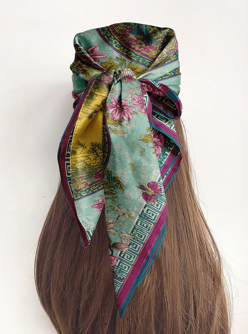 May include: A floral patterned headscarf tied around the head. The scarf features a light blue background with pink flowers and a yellow accent. The edges are trimmed with a dark purple border. The scarf is wrapped around the hair.