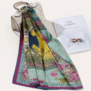 May include: A colorful scarf with a floral and architectural design, draped over a white handbag. The scarf features shades of teal, yellow, and pink with a geometric border. A book with the words "LOVE FLOWERS" is in the background.
