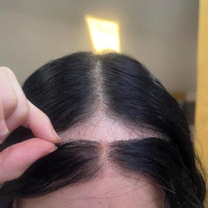 May include: Close-up of a black wig with a lace front. The wig's hair is parted in the middle, and the lace front is visible. The hair is dark and appears to be styled.
