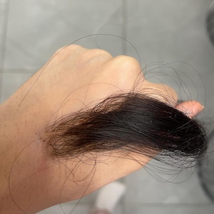 Può includere: Una mano che tiene in mano una ciocca di capelli castano scuro. I capelli sono di lunghezze variabili, con alcune ciocche più lunghe di altre. I capelli sembrano appena tagliati.