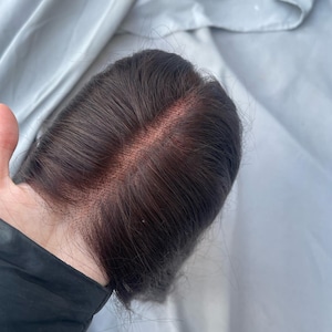 Könnte beinhalten: Ein dunkelbraunes Haarteil mit Spitzenfront, das einen sichtbaren Scheitel aufweist. Das Haar ist mit einer natürlichen Welle versehen. Das Haarteil liegt vor einem hellgrauen Stoffhintergrund.