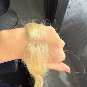 May include: Blonde hair extensions, a bundle of light-colored hair, held in a hand. The hair is a bright, pale blonde color, with a slightly wavy texture. Strands of hair are visible.