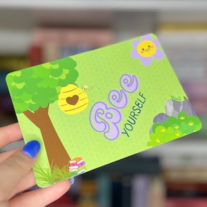 May include: A rectangular card with a green background featuring a tree, a yellow beehive with a heart, and the words "Bee Yourself" in purple. A smiling flower and small rocks are also depicted.