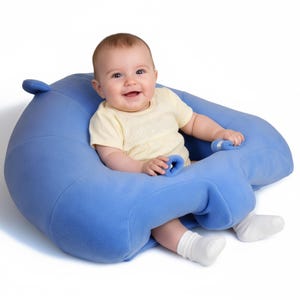 Puede incluir: Un bebé está sentado en un asiento para bebé de felpa azul claro. El asiento tiene forma de hipopótamo con respaldo curvo y reposabrazos. El bebé lleva una camisa amarilla y calcetines blancos, sonriendo a la cámara.