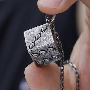 May include: A silver-colored pendant in the shape of a die, featuring multiple eye-shaped blue gemstones. The pendant is suspended from a silver chain. The die is held between fingers.