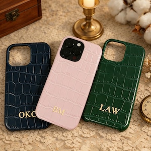 May include: Three phone cases in navy, pink, and green, each with a crocodile skin texture. The navy case has "OKO" in gold lettering, the pink case has "BM", and the green case has "LAW".