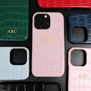 Puede incluir: Una colección de fundas para teléfono de varios colores con textura de piel de cocodrilo. Las fundas son de color verde, rojo, azul, rosa y negro. Cada funda tiene las letras "ABC" en dorado. Diseñadas para smartphones.