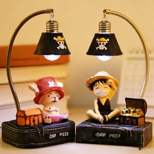 May include: Two decorative lamps featuring cartoon characters. Each lamp has a black lampshade with a skull and crossbones design. The lamps are supported by flexible metal arms. One lamp features a character in a red hat, the other in a straw hat. The bases read "ONE PIECE".