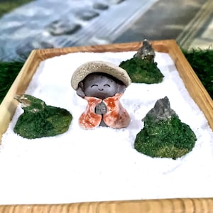 Może przedstawiać: Miniaturowy ogród zen w drewnianej tacy. Ogród przedstawia uśmiechniętą figurkę Buddy w kapeluszu i szacie, otoczoną małymi formacjami skalnymi pokrytymi mchem na białym piasku. Taca jest prostokątna z naturalnym wykończeniem drewna.