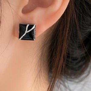 May include: A square stud earring featuring a glossy black surface with a thin, white, Y-shaped line design. The earring is secured to the earlobe, showcasing a smooth, polished finish and a geometric shape that contrasts with the white accent line.