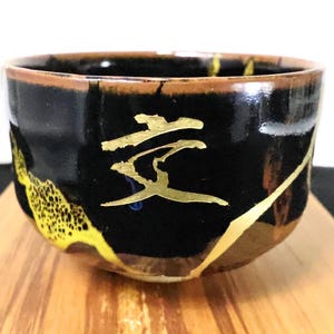 Può includere: Una ciotola da tè in ceramica nera con un carattere giapponese dorato sulla parte anteriore. La ciotola presenta un bordo marrone e accenti decorativi dorati con un motivo testurizzato e maculato sul lato inferiore, rifinito con smalto lucido.