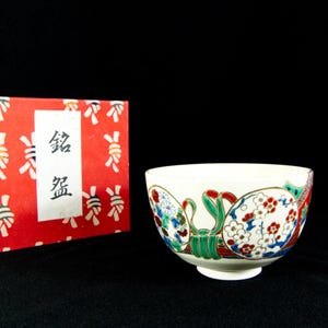 Könnte beinhalten: Eine weiße Keramikschale mit einem dekorativen Blumendesign in Rot, Blau, Grün und Gold. Die Schale befindet sich neben einer roten Schachtel mit japanischem Text und weißen und goldenen Mustern. Die Schale hat einen kleinen Fuß.