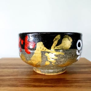 Pode incluir: Uma tigela de cerâmica com acabamento texturizado, apresentando uma seção superior preta e uma seção inferior cinza salpicada. O exterior é decorado com formas abstratas douradas pintadas à mão, detalhes em vermelho e texto branco à direita.