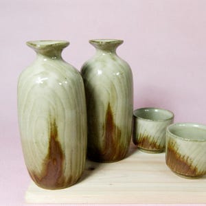 Könnte beinhalten: Ein Keramik-Sake-Set mit zwei hohen, hellgrünen Flaschen mit braunen Akzenten und zwei passenden Tassen. Das Set wird auf einer hellen Holzoberfläche vor einem zartrosa Hintergrund präsentiert. Die Flaschen und Tassen haben ein rustikales, handgefertigtes Aussehen.