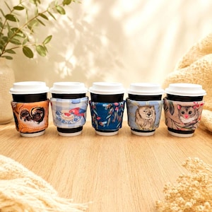 May include: Five reusable fabric coffee cup sleeves featuring diverse illustrations including two small animals, a bird on a plate, eucalyptus leaves, a wombat, and a possum. Each sleeve is wrapped around a black cup with a white lid.
