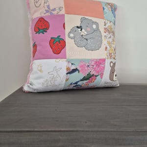 May include: A patchwork cushion with various patterns and images. The cushion features squares with strawberries, butterflies, a koala bear with the text "MAMA BEAR AND ME", and floral designs. The cushion has a pink trim.