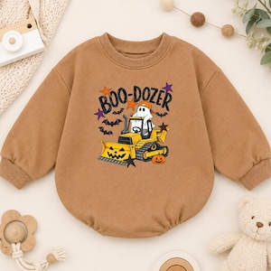 May include: A tan long-sleeve sweatshirt featuring a graphic print of a ghost wearing an orange cap driving a yellow bulldozer. The bulldozer blade is shaped like a jack-o'-lantern. The text "BOO-DOZER" is printed above, surrounded by bats and stars.