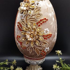 May include: A decorative egg-shaped ornament with a beige velvet surface, adorned with gold-colored floral embellishments, red and clear crystals, and white pearls. The egg rests on a decorative stand with a silver and gold base, set against a black background.