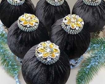 Puede incluir: Seis adornos navideños de terciopelo negro con adornos florales con incrustaciones de joyas. Cada adorno se asienta sobre una base decorativa plateada. Los adornos están dispuestos sobre una superficie clara, con un collar de perlas y una ramita verde.