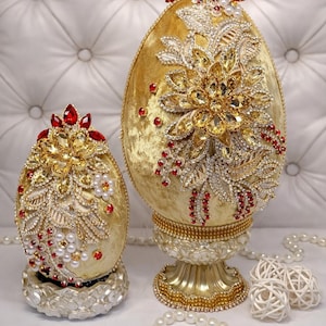 Pode incluir: Dois ornamentos decorativos de ovo dourado. Os ovos são adornados com intrincados desenhos florais feitos de cristais, pérolas e pedras preciosas vermelhas. Um ovo assenta sobre um pedestal, enquanto o outro repousa sobre uma base decorativa. Uma fileira de pérolas e bolas decorativas também estão presentes.