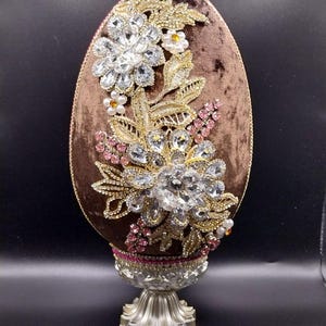 Pode incluir: Um ovo decorativo de veludo castanho adornado com bordados dourados, cristais transparentes e detalhes rosa e branco. O ovo assenta numa base de pedestal prateada, criando uma peça de exibição elegante. O ovo é um item decorativo único.