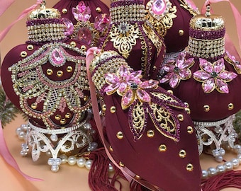 May include: A group of decorative burgundy ornaments with gold and rhinestone detailing. Each has pink gemstones, tassels, and is displayed on decorative stands with pearl accents and pink ribbons. The ornaments are highly embellished.