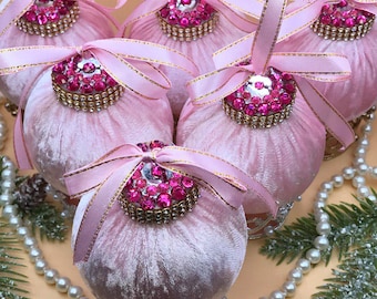 May include: Six pink velvet Christmas baubles with jeweled tops and pink ribbon bows. The baubles are decorated with gold trim and sit on decorative silver bases. A string of pearls and pine branches are in the background.