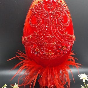 Op de afbeelding: Een decoratief rood, eivormig ornament. Het is versierd met rode kralen en strass, met een bloemmotief en een basis van rode veren. Het ei is felrood, tegen een donkere achtergrond. Afmetingen in centimeters.