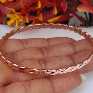 May include: A copper-colored bracelet with a braided design. The bracelet is circular and appears to be made of interwoven metal strands. The image shows the bracelet resting on a hand, with the text "Ryanshijewellers" visible.