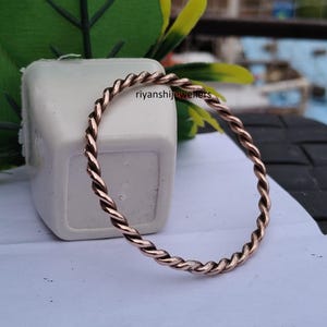 May include: A copper-colored twisted bangle bracelet. The bracelet has a rope-like design and is displayed on a white surface. The text "riyanshijewellers" is visible on the background.