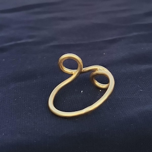 Op de afbeelding: Een goudkleurig, abstract gevormd sieraad. Het ontwerp heeft een lusvormige bovenkant en een gebogen, langwerpige onderkant, waardoor een unieke, vloeiende vorm ontstaat. Het sieraad staat tegen een donkerblauwe achtergrond.