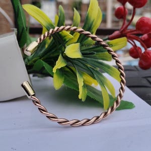May include: A copper-colored twisted metal bracelet with gold-colored end caps. The bracelet is open and sits on a white surface, with green and yellow leaves and red berries in the background. The text "Ryansjewellers" is visible.