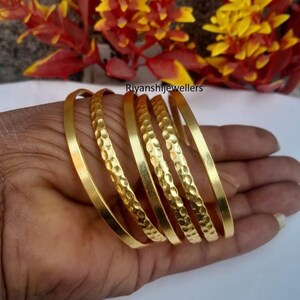 Op de afbeelding: Een hand toont een verzameling gouden armbanden. Sommige armbanden hebben een glad, gepolijst oppervlak, terwijl andere een getextureerd, gehamerd patroon hebben. De sieraden worden gepresenteerd tegen een achtergrond van kleurrijke bloemen.
