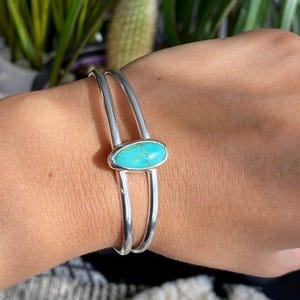 Peut inclure: Un bracelet manchette argenté avec une pierre ovale turquoise vif au centre. Le bracelet a un design à double bande, la pierre étant sertie dans un sertissage à lunette. Le bracelet est porté sur un poignet.