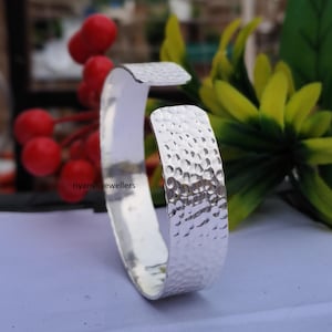 May include: A silver cuff bracelet with a hammered texture. The open-ended design features a rectangular shape with rounded edges. The bracelet is displayed against a backdrop of red berries and green foliage.