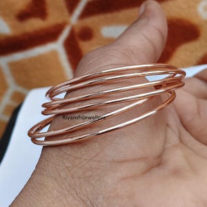 Op de afbeelding: Een stapel roségouden armbanden. De dunne, ronde armbanden zijn op de hand van een persoon gestapeld. De armbanden hebben een glanzende, metalen afwerking. De merknaam "Riyanshijewellers" is zichtbaar.