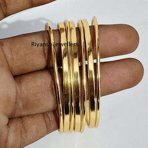May include: A collection of gold-coloured bangles, stacked together, with a smooth, reflective surface. The bangles are held in a hand, with the text "Ryanshijewellers" visible. The bangles are a classic jewellery item.