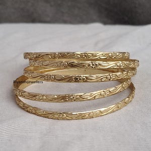 Peut inclure: Plusieurs bracelets dorés avec un motif floral. Les bracelets sont empilés, mettant en valeur le motif floral sur leur surface. Les bijoux sont présentés sur une surface blanche.