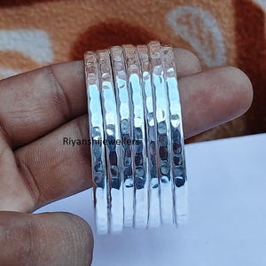 May include: A collection of silver rings with a hammered finish. The rings are shown against a blurred backdrop. The rings are made of a shiny, reflective metal.