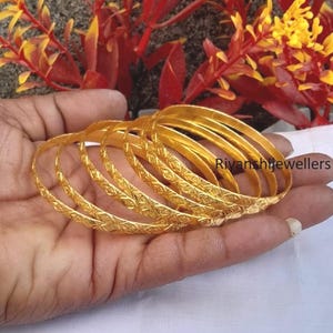 Puede incluir: Un conjunto de pulseras doradas con un patrón detallado y repetitivo. Las pulseras están apiladas y la imagen muestra una vista de cerca de las joyas. El texto "Rivanshi jewellers" es visible en la imagen.