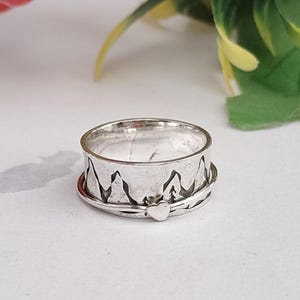 Peut inclure: Une bague argentée ornée d'un motif de chaîne de montagnes sur le pourtour. Un petit cœur est attaché à la bague. La bague est posée sur une surface blanche.