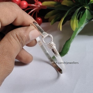 May include: A silver bangle bracelet with a simple design. The bracelet has a rounded shape and a clasp closure. The text "Riyanshijewellers" is visible on the bracelet.
