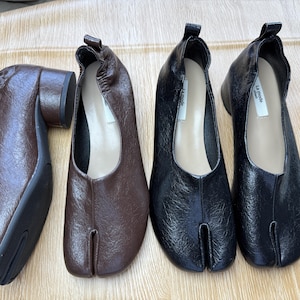 Op de afbeelding: Vier paar leren schoenen in bruin- en zwarttinten. De schoenen hebben een split-teenontwerp en een lage hak. De schoenen zijn gerangschikt op een lichtgekleurd houten oppervlak.