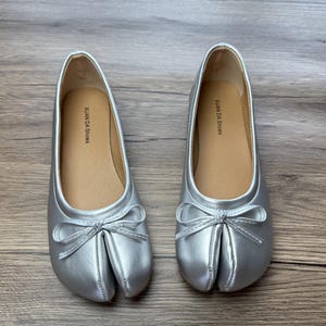 May include: A pair of silver ballet flats with a split-toe design and a small bow detail. The shoes have a shiny, metallic finish and a tan insole with the text "XUAN DA Shoes".