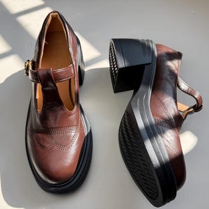 Könnte beinhalten: Paar braune Mary Jane Plateauschuhe aus Leder. Die Schuhe haben einen T-Riemen mit Schnalle, eine klobige schwarze Sohle und einen Absatz sowie dekorative Nähte. Absatzhöhe ca. 7,6 cm.